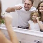 family brushing their teeth together in bathroom mirror